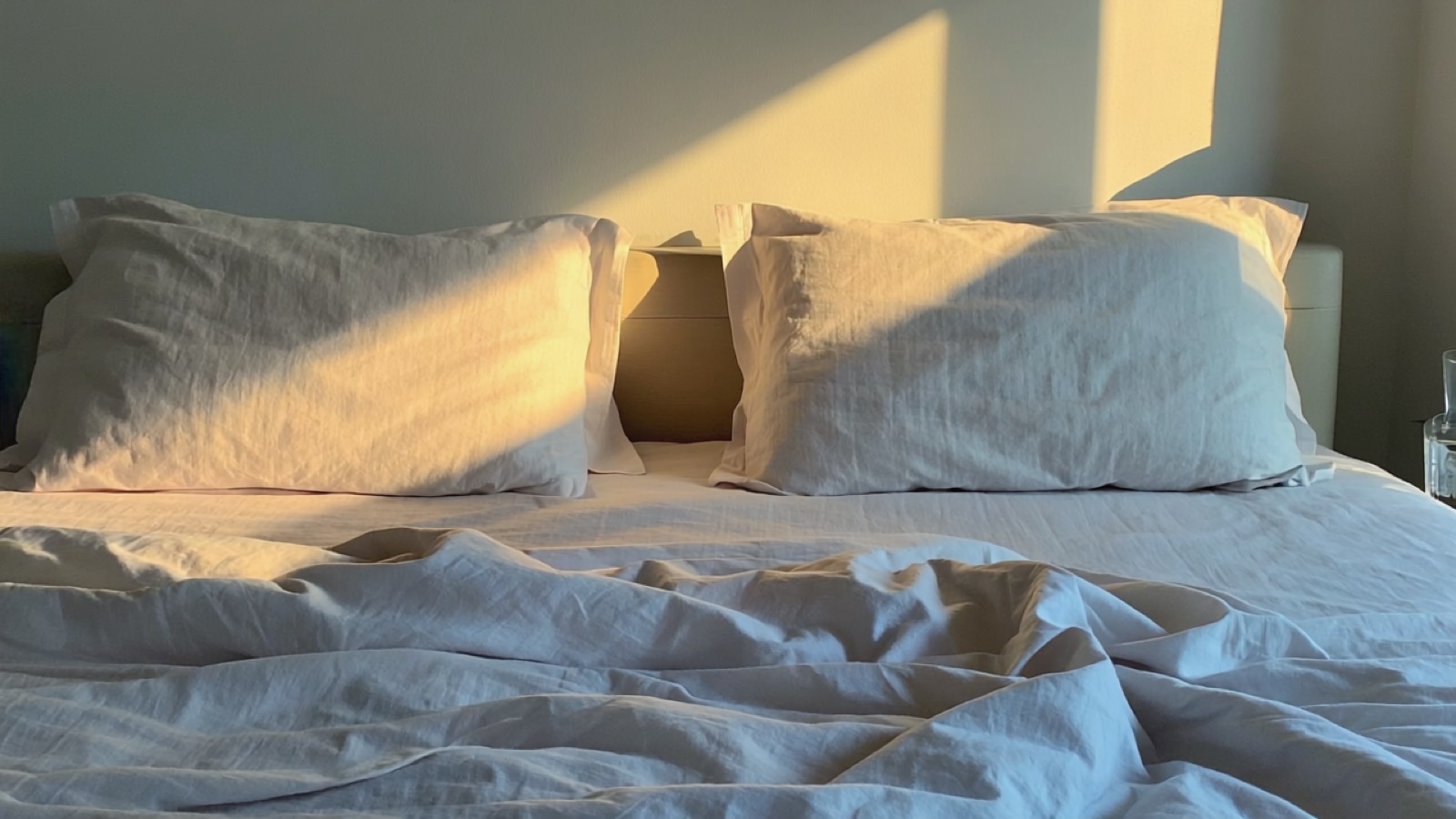 Zerknitterte Leinenbettwäsche im blassen Morgenlicht, ein ruhiges Schlafzimmer mit einem Glas Wasser auf dem Nachttisch